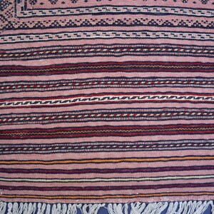 May include: A woven textile rug featuring a dusty pink base color with horizontal stripes in dark blue, deep red, and cream. The pattern includes intricate geometric borders and a series of thin, repeating lines, finished with a white fringe edge.