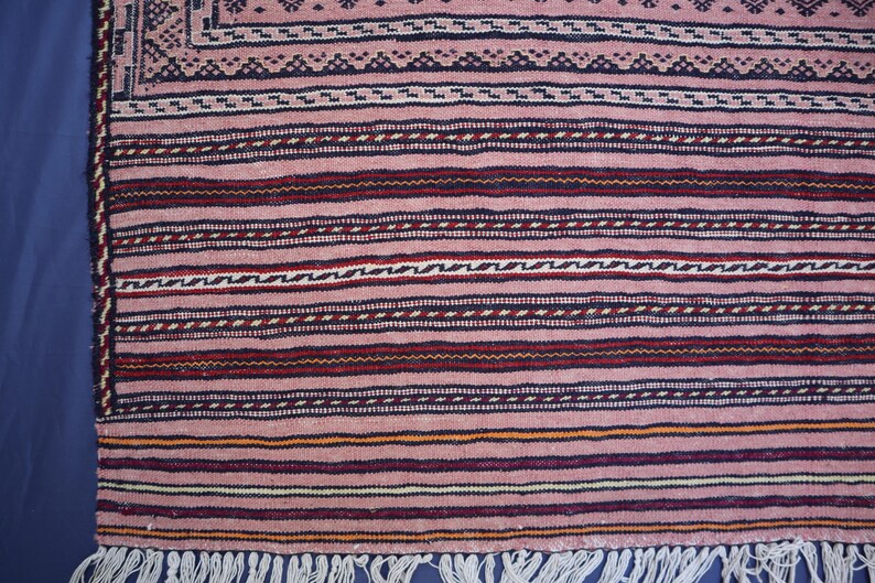 May include: A woven textile rug featuring a dusty pink base color with horizontal stripes in dark blue, deep red, and cream. The pattern includes intricate geometric borders and a series of thin, repeating lines, finished with a white fringe edge.