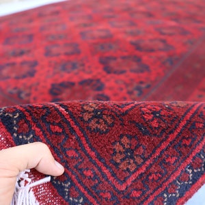 May include: A red and black patterned rug with fringe. The rug is folded in half and being held by a hand.