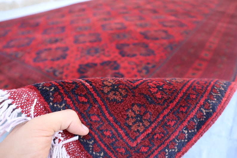 May include: A red and black patterned rug with fringe. The rug is folded in half and being held by a hand.