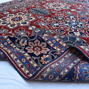 K&ouml;nnte beinhalten: Ein rechteckiger Teppich mit tiefrotem Hintergrund und komplexen Blumen- und Geometriemustern in Marineblau, Creme und Hellblau. Der Teppich ist umgeschlagen und zeigt einen dunkelblauen Rand mit sich wiederholenden Stern- und Blumenmotiven.