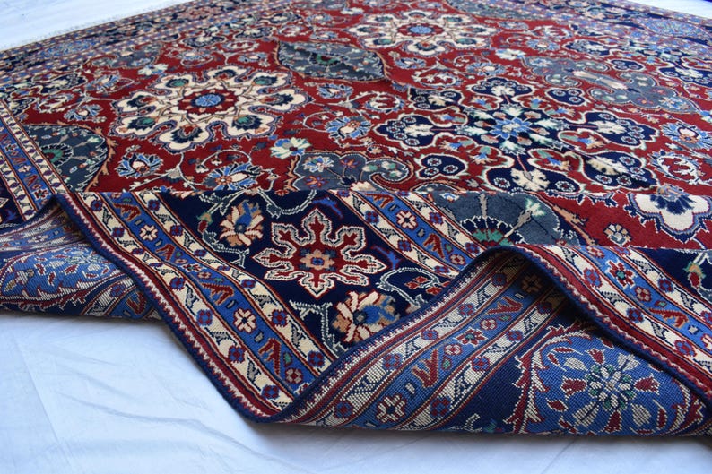 K&ouml;nnte beinhalten: Ein rechteckiger Teppich mit tiefrotem Hintergrund und komplexen Blumen- und Geometriemustern in Marineblau, Creme und Hellblau. Der Teppich ist umgeschlagen und zeigt einen dunkelblauen Rand mit sich wiederholenden Stern- und Blumenmotiven.