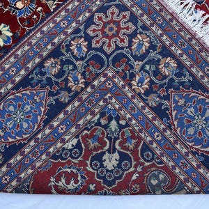 K&ouml;nnte beinhalten: Ein gewebter Teppich mit einem tiefblauen Mittelfeld, das komplexe florale und geometrische Muster in Rot, Hellblau und Creme aufweist. Der Rand zeigt einen dunkelroten Hintergrund mit &auml;hnlichen Blumenmotiven und wei&szlig;en Fransen.