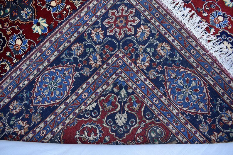 K&ouml;nnte beinhalten: Ein gewebter Teppich mit einem tiefblauen Mittelfeld, das komplexe florale und geometrische Muster in Rot, Hellblau und Creme aufweist. Der Rand zeigt einen dunkelroten Hintergrund mit &auml;hnlichen Blumenmotiven und wei&szlig;en Fransen.