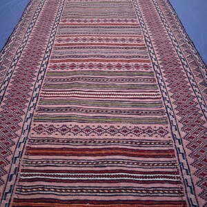 May include: A long, rectangular runner rug featuring a repeating pattern of geometric diamonds and horizontal stripes in shades of dusty pink, deep red, dark blue, and cream. The border displays a continuous zigzag motif in dark blue and pink.