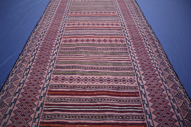May include: A long, rectangular runner rug featuring a repeating pattern of geometric diamonds and horizontal stripes in shades of dusty pink, deep red, dark blue, and cream. The border displays a continuous zigzag motif in dark blue and pink.