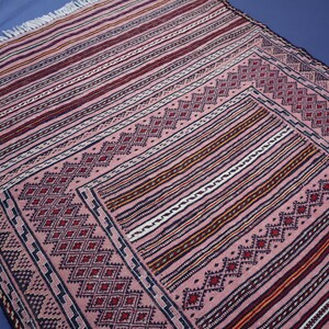 May include: A rectangular woven rug featuring a dusty pink base with intricate geometric diamond patterns in dark red and black. The design includes horizontal striped borders with white fringe at the top edge, showcasing a detailed textile construction.