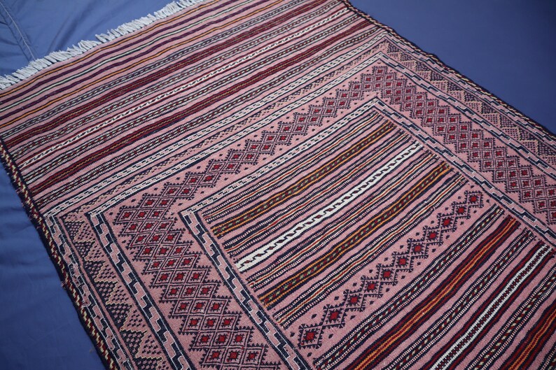 May include: A rectangular woven rug featuring a dusty pink base with intricate geometric diamond patterns in dark red and black. The design includes horizontal striped borders with white fringe at the top edge, showcasing a detailed textile construction.