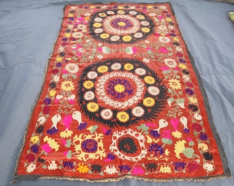 May include: A rectangular, red textile with intricate floral and geometric patterns. The design features large circular motifs with concentric rings of flowers in yellow, white, and purple. The edges are bordered with a dark gray trim. The textile is likely a decorative piece.