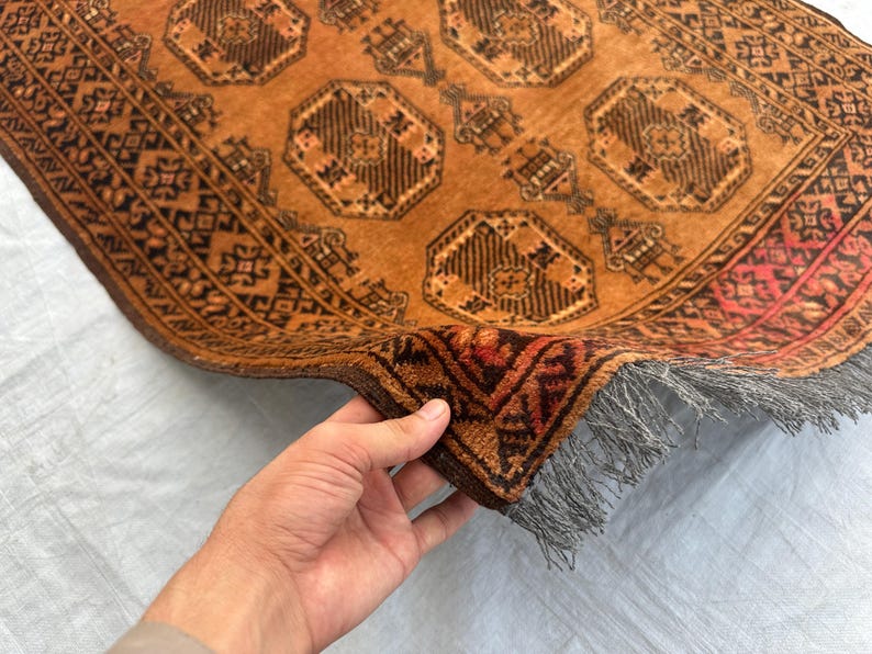 May include: A rectangular rug featuring a warm orange background with repeating dark brown octagonal geometric patterns and intricate border designs. The rug has a thick pile texture and includes a short, light gray fringe along the edge.