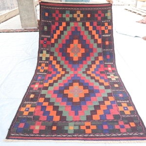 May include: A colorful geometric rug with a repeating pattern of squares and diamonds in shades of red, orange, green, blue, and brown. The rug has a fringe border.