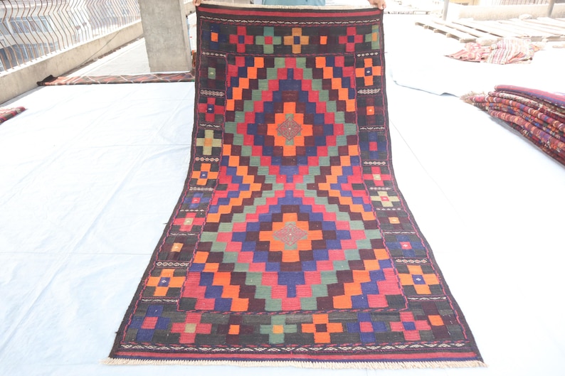 May include: A colorful geometric rug with a repeating pattern of squares and diamonds in shades of red, orange, green, blue, and brown. The rug has a fringe border.