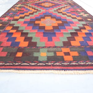 May include: A colorful geometric rug with a traditional design. The rug features a variety of colors, including red, orange, green, blue, and brown. The rug has a fringe border.