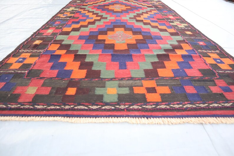 May include: A colorful geometric rug with a traditional design. The rug features a variety of colors, including red, orange, green, blue, and brown. The rug has a fringe border.
