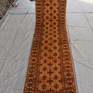 Puede incluir: Una alfombra de pasillo larga y estrecha con un campo de color amarillo dorado y un patrón repetitivo de motivos de cruces geométricas y bordes cuadrados en marrón oscuro. La alfombra tiene una banda horizontal roja oscura.
