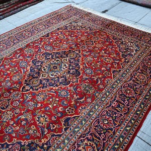 Puede incluir: Una alfombra rectangular grande con un fondo rojo vibrante y un medall&oacute;n central intrincado en azul oscuro, crema y dorado. La alfombra est&aacute; decorada con patrones detallados de flores y enredaderas, terminada con flecos blancos.