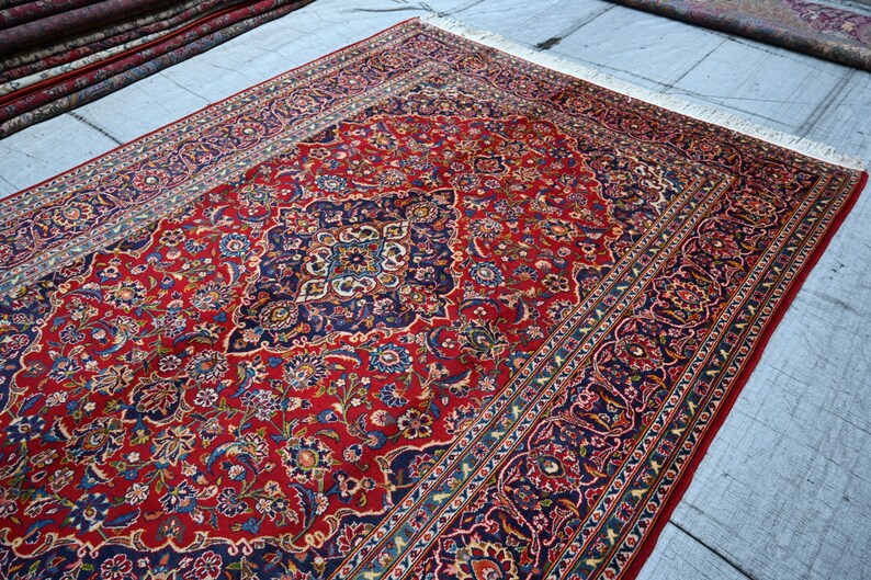 Puede incluir: Una alfombra rectangular grande con un fondo rojo vibrante y un medall&oacute;n central intrincado en azul oscuro, crema y dorado. La alfombra est&aacute; decorada con patrones detallados de flores y enredaderas, terminada con flecos blancos.