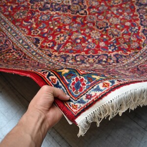 Puede incluir: Una alfombra rectangular con un denso patr&oacute;n floral y de cachemira en tonos rojo, azul marino y crema. La alfombra tiene un borde decorativo ancho con motivos intrincados y un fleco de borlas blancas a lo largo del borde corto del tejido.