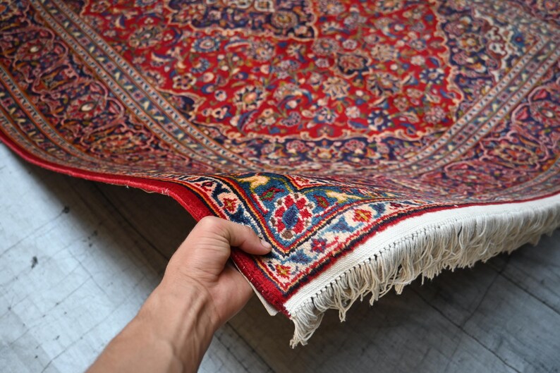 Puede incluir: Una alfombra rectangular con un denso patr&oacute;n floral y de cachemira en tonos rojo, azul marino y crema. La alfombra tiene un borde decorativo ancho con motivos intrincados y un fleco de borlas blancas a lo largo del borde corto del tejido.