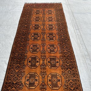 May include: A rectangular runner rug featuring a vibrant burnt orange background with repeating dark brown octagonal geometric motifs arranged in two vertical columns. The border displays intricate black and brown patterns with fringe ends.