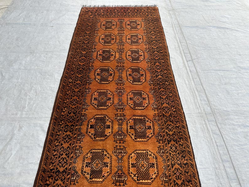 May include: A rectangular runner rug featuring a vibrant burnt orange background with repeating dark brown octagonal geometric motifs arranged in two vertical columns. The border displays intricate black and brown patterns with fringe ends.