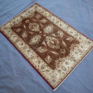 May include: Rectangular brown rug with a floral pattern and a cream-colored border. The rug has a red edge and is placed on a blue surface. The rug's design features a central field of brown with cream-colored floral motifs.