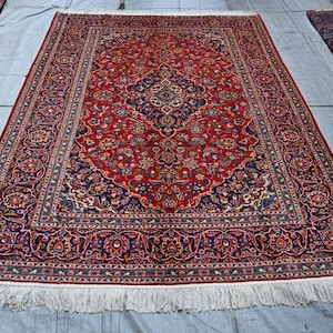 Puede incluir: Una alfombra rectangular que presenta un campo rojo vibrante con un medall&oacute;n central azul oscuro y patrones florales intrincados en tonos azul, crema y dorado. La alfombra tiene un borde decorativo ancho y flecos blancos en los extremos.