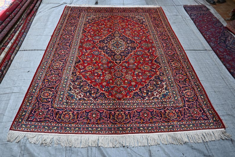 Puede incluir: Una alfombra rectangular que presenta un campo rojo vibrante con un medall&oacute;n central azul oscuro y patrones florales intrincados en tonos azul, crema y dorado. La alfombra tiene un borde decorativo ancho y flecos blancos en los extremos.