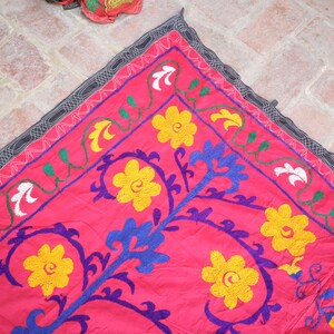 K&ouml;nnte beinhalten: Ein leuchtend pinkfarbener Stoff mit aufwendiger Stickerei. Er zeigt gelbe, wei&szlig;e und blaue Blumenmuster und gr&uuml;ne Ranken. Der Rand ist dunkelgrau mit einem sich wiederholenden Muster. Der Stoff ist wahrscheinlich ein dekoratives St&uuml;ck.