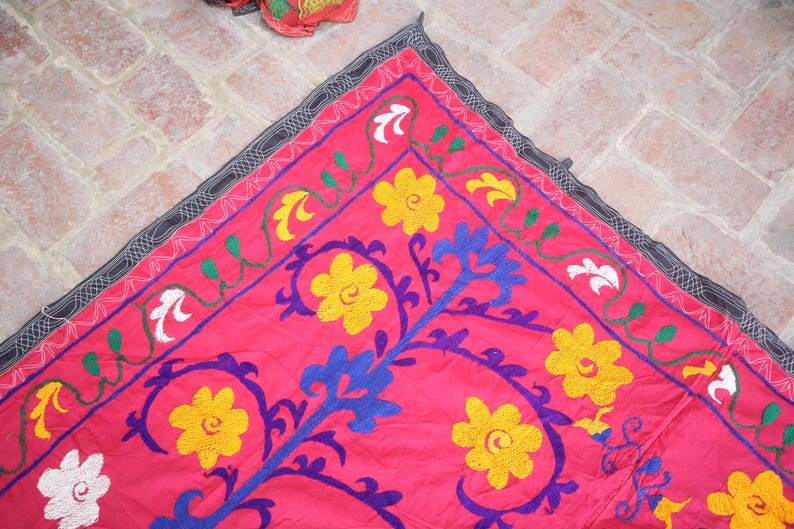 K&ouml;nnte beinhalten: Ein leuchtend pinkfarbener Stoff mit aufwendiger Stickerei. Er zeigt gelbe, wei&szlig;e und blaue Blumenmuster und gr&uuml;ne Ranken. Der Rand ist dunkelgrau mit einem sich wiederholenden Muster. Der Stoff ist wahrscheinlich ein dekoratives St&uuml;ck.