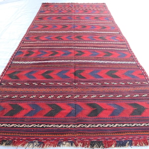 Puede incluir: Una alfombra roja, azul y negra con un borde de flecos. La alfombra tiene un diseño geométrico con formas de flecha repetidas.