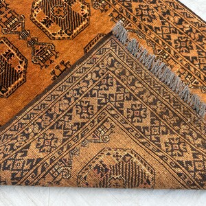 May include: A hand-knotted rug featuring a deep orange background with repeating black geometric octagonal motifs and intricate border patterns. The rug is folded over to reveal the underside, showcasing a dense weave and short grey fringe tassels.