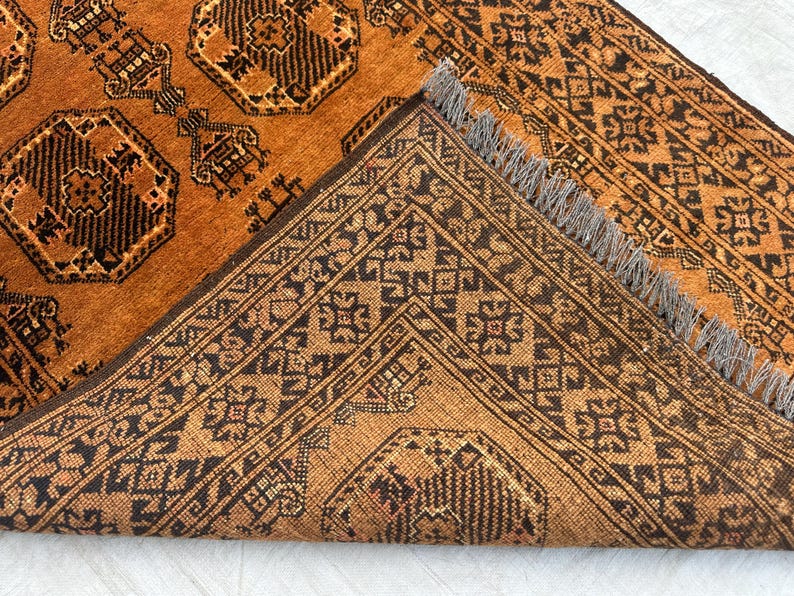 May include: A hand-knotted rug featuring a deep orange background with repeating black geometric octagonal motifs and intricate border patterns. The rug is folded over to reveal the underside, showcasing a dense weave and short grey fringe tassels.