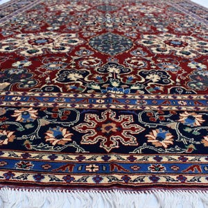 K&ouml;nnte beinhalten: Ein gro&szlig;er rechteckiger Teppich mit einem tiefroten Mittelfeld, das mit komplizierten Blumen- und Medaillonmustern in Marineblau, Creme und Hellblau verziert ist. Der Teppich hat eine breite marineblaue Bord&uuml;re und wei&szlig;e Fransen am Rand.