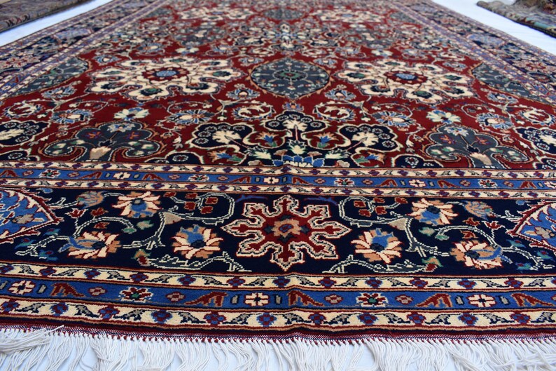 K&ouml;nnte beinhalten: Ein gro&szlig;er rechteckiger Teppich mit einem tiefroten Mittelfeld, das mit komplizierten Blumen- und Medaillonmustern in Marineblau, Creme und Hellblau verziert ist. Der Teppich hat eine breite marineblaue Bord&uuml;re und wei&szlig;e Fransen am Rand.