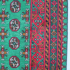 K&ouml;nnte beinhalten: Ein gr&uuml;ner Teppich mit einem roten, braunen und gr&uuml;nen Rand. Der Teppich hat ein sich wiederholendes Muster aus geometrischen Formen.
