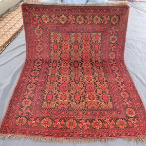 May include: A rectangular, hand-woven rug with a detailed floral pattern in shades of red, black, and gold. The rug features a central medallion design and a decorative border. The rug has a fringed edge.