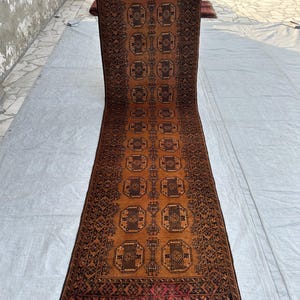May include: A long, rectangular runner rug featuring a warm, burnt orange background with repeating dark brown octagonal geometric motifs. The rug is framed by a detailed dark brown border and finished with short, grey fringe at both ends.