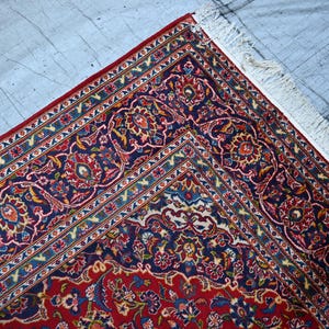 Puede incluir: Una alfombra rectangular con un campo central de color rojo intenso y un borde ancho de color azul marino, ambos decorados con intrincados patrones florales y de enredaderas en tonos azul claro, crema y dorado. La alfombra tiene flecos blancos.