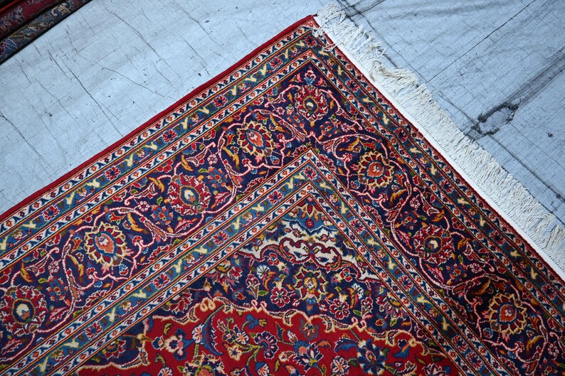 Puede incluir: Una alfombra rectangular con un campo central de color rojo intenso y un borde ancho de color azul marino, ambos decorados con intrincados patrones florales y de enredaderas en tonos azul claro, crema y dorado. La alfombra tiene flecos blancos.