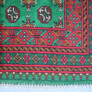 K&ouml;nnte beinhalten: Ein gr&uuml;ner, roter und brauner Teppich mit Fransenrand. Der Teppich hat ein geometrisches Muster mit sich wiederholenden Motiven.