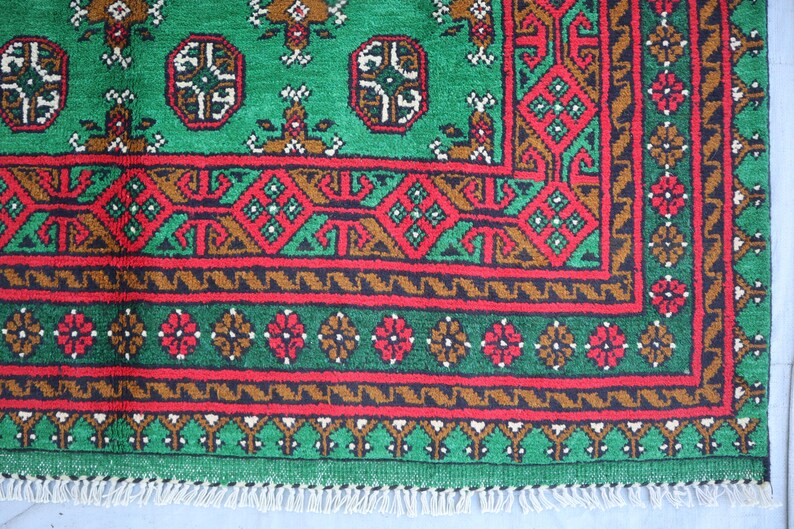 K&ouml;nnte beinhalten: Ein gr&uuml;ner, roter und brauner Teppich mit Fransenrand. Der Teppich hat ein geometrisches Muster mit sich wiederholenden Motiven.