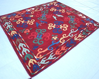 May include: A red, square rug with a colorful, intricate design. The rug features a repeating pattern of stylized flowers, birds, and swirls. The border of the rug is a repeating pattern of stylized birds and swirls.