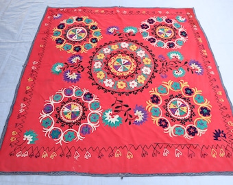May include: A red fabric square with intricate embroidery in various colors, including white, black, green, yellow, and purple. The embroidery features multiple circular designs with floral motifs.