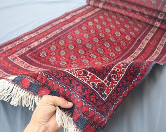 Könnte beinhalten: Ein rot-blauer Teppich mit Muster und Fransenrand. Der Teppich ist in der Mitte gefaltet und wird von einer Hand gehalten.