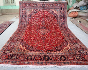 May include: A large, rectangular red rug with an intricate floral pattern and a dark blue border. The rug has a central medallion design and a cream-coloured background. The rug is displayed on a light-coloured surface.