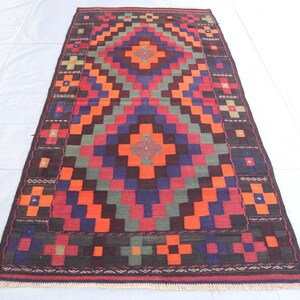 May include: A traditional rug with a geometric pattern in shades of red, orange, blue, green, and brown. The rug has a central diamond shape with a smaller diamond inside. The rug is surrounded by a border with a repeating pattern.