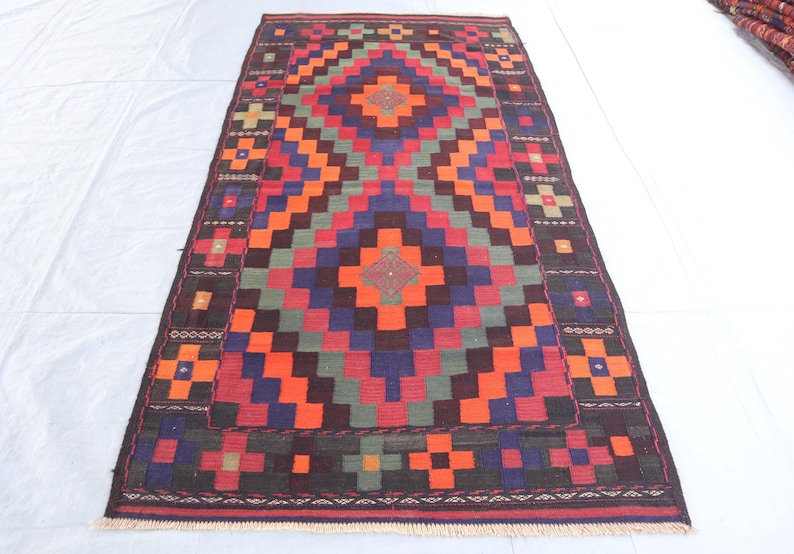May include: A traditional rug with a geometric pattern in shades of red, orange, blue, green, and brown. The rug has a central diamond shape with a smaller diamond inside. The rug is surrounded by a border with a repeating pattern.