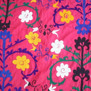 K&ouml;nnte beinhalten: Nahaufnahme eines leuchtend rosa Textils mit gestickten Blumenmustern. Das Design zeigt wei&szlig;e, gelbe und schwarze Blumen sowie blaue und gr&uuml;ne Akzente. Der Stoff hat ein strukturiertes Aussehen und zeigt die detaillierte Handarbeit.
