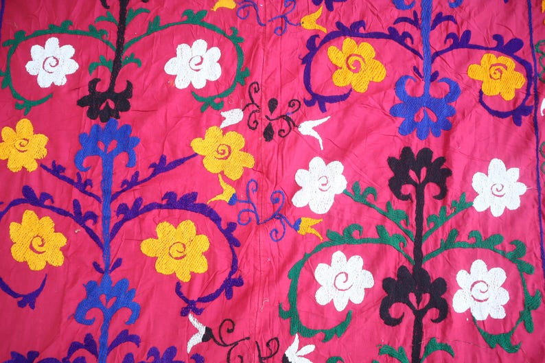K&ouml;nnte beinhalten: Nahaufnahme eines leuchtend rosa Textils mit gestickten Blumenmustern. Das Design zeigt wei&szlig;e, gelbe und schwarze Blumen sowie blaue und gr&uuml;ne Akzente. Der Stoff hat ein strukturiertes Aussehen und zeigt die detaillierte Handarbeit.